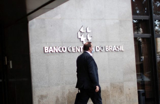 Homem de terno caminha em frente ao Banco Central do Brasil, com chão molhado e reflexivo.