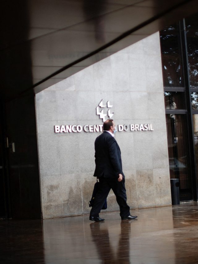 Homem de terno caminha em frente ao Banco Central do Brasil, com chão molhado e reflexivo.