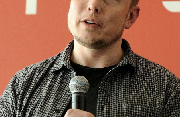 Elon Musk, em camisa xadrez, fala ao microfone. Ao fundo, o logo da Tesla em uma parede vermelha.