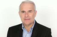 Retrato profissional de homem com cabelos grisalhos, blazer preto e camisa listrada em fundo branco.