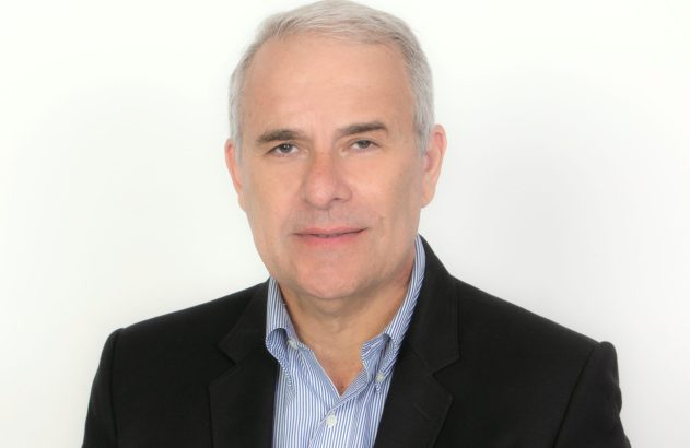 Retrato profissional de homem com cabelos grisalhos, blazer preto e camisa listrada em fundo branco.