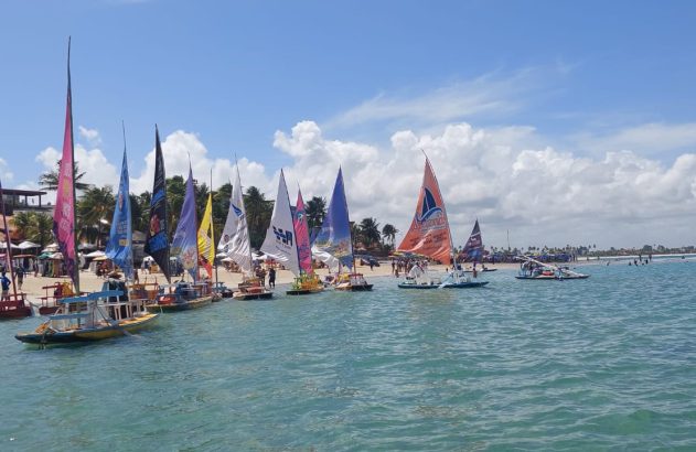 Praia tropical ensolarada com muitas jangadas de velas coloridas, pessoas na areia e no mar, e palmeiras ao fundo.