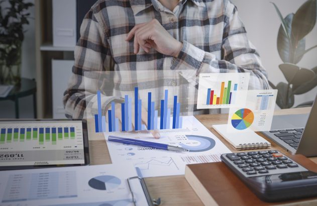 Pessoa analisando gráficos financeiros holográficos e documentos em uma mesa de escritório.