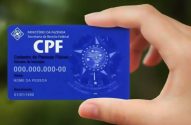 Mão segura cartão CPF azul brasileiro com nome e data de nascimento fictícios.