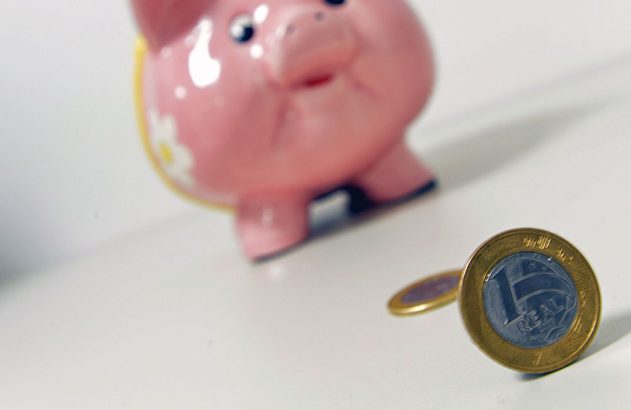 Cofrinho rosa e moedas de Real. Uma moeda de 1 Real está em destaque, simbolizando poupança.