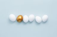 Um ovo dourado alinhado com cinco ovos brancos em um fundo azul claro.