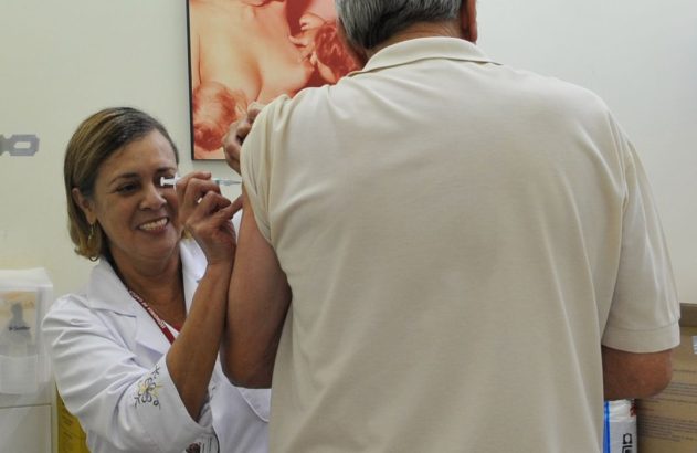 Profissional de saúde sorridente aplica vacina no braço de um paciente.