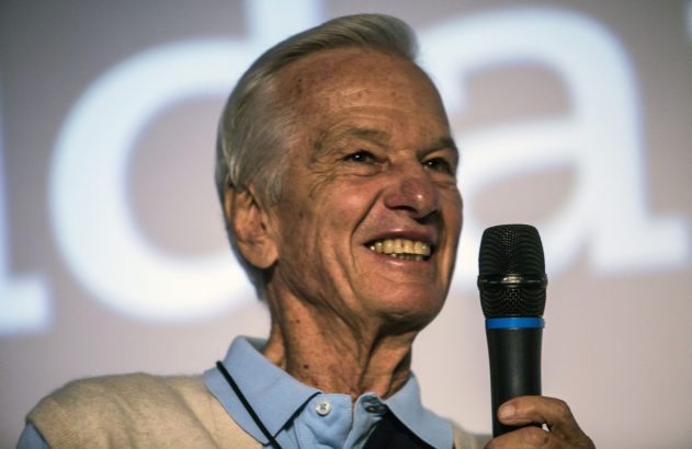 Homem idoso sorridente, de cabelo grisalho, segurando um microfone. No fundo desfocado, letras grandes em branco.
