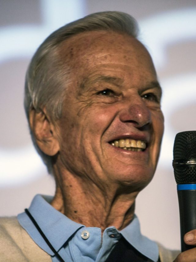 Homem idoso sorridente, de cabelo grisalho, segurando um microfone. No fundo desfocado, letras grandes em branco.