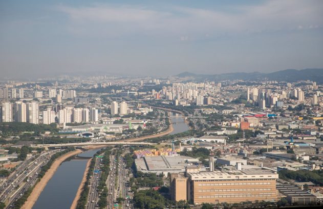 Vista aérea de uma metrópole com rio, rodovias com tráfego intenso e muitos edifícios. Montanhas ao fundo.