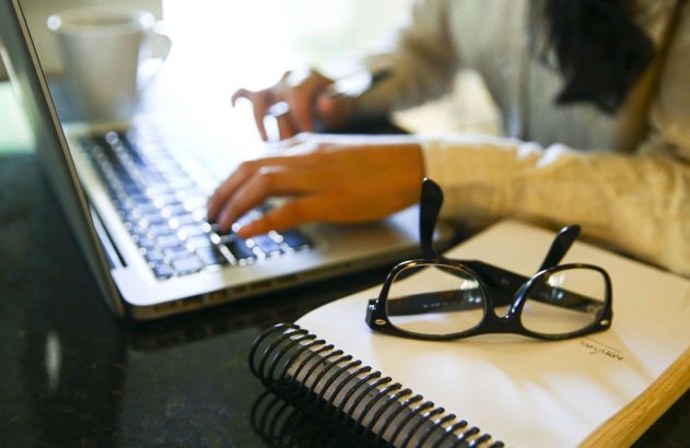Mãos digitando em laptop. Óculos e caderno sobre mesa escura.