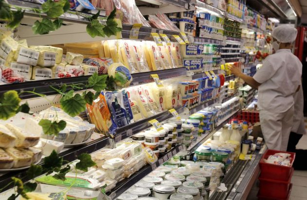 Pessoa repõe queijos e laticínios em prateleiras refrigeradas de supermercado, com decoração de folhas.