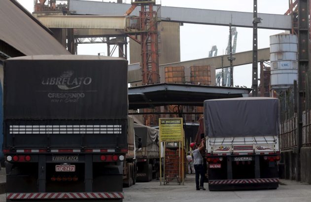Caminhões de carga estacionados em pátio industrial. Um homem ajusta lona de caminhão, com silos e guindastes ao fundo.