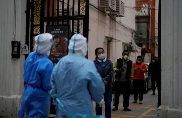 Profissionais em EPI azul e civis com máscaras em entrada de prédio, durante prevenção à pandemia em 2022.