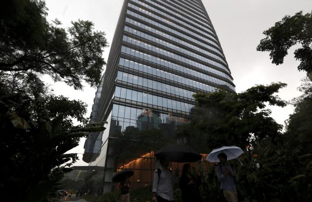 Pessoas com guarda-chuvas caminham em frente a um arranha-céu moderno, cercado por árvores, sob céu nublado.