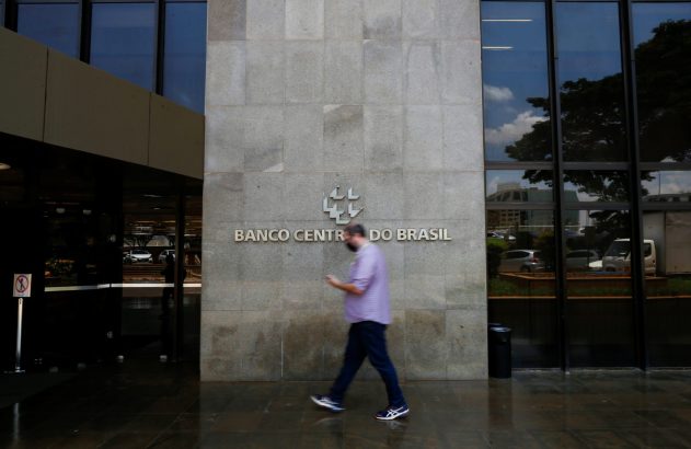 Homem desfocado, olhando o celular, passa em frente ao prédio do Banco Central do Brasil com janelas de vidro.