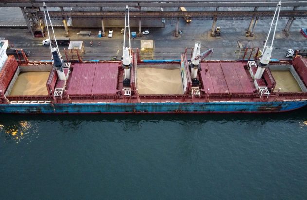 Vista aérea de navio graneleiro em porto, com porões abertos e parcialmente carregados com grãos.