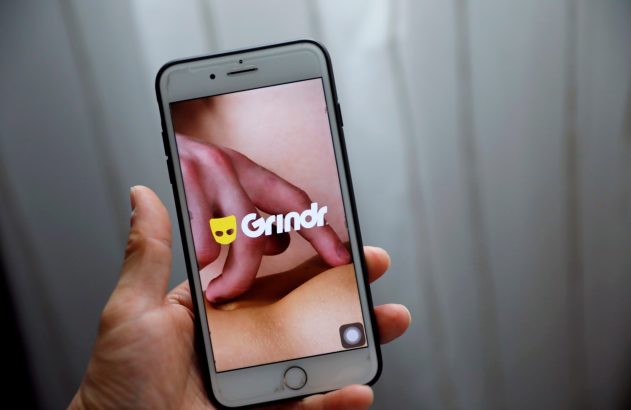 Mão segura smartphone. Tela exibe logo do Grindr sobre close-up de pele com dedos.