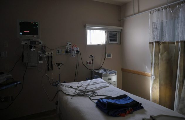 Quarto de hospital com cama desocupada, equipamentos médicos na parede, janela e cortinas.