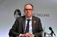 Homem de terno, óculos e papoula, sentado. Ao fundo, logo e nome do Banco da Inglaterra, durante coletiva de imprensa.