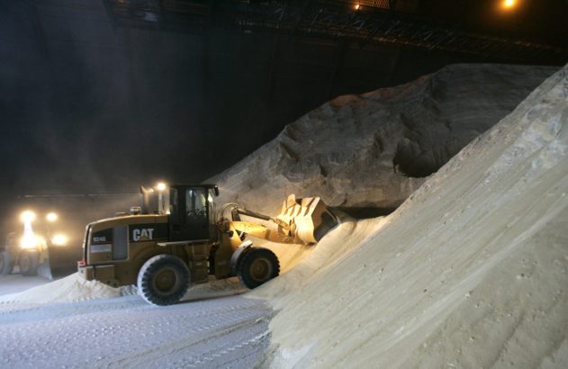 Carregadeira CAT amarela em armazém escuro, empurrando uma vasta pilha de material granular claro.