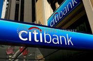 Letreiro azul e branco da Citibank em prédio, com reflexo de pessoas e outros edifícios ao fundo.