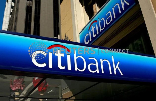 Letreiro azul e branco da Citibank em prédio, com reflexo de pessoas e outros edifícios ao fundo.