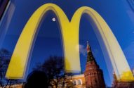 Logo amarelo do McDonald's. Refletida em vidro, uma torre do Kremlin de Moscou sob céu azul.