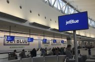 Interior de terminal de aeroporto JetBlue com pessoas em balcões de atendimento e telões exibindo a marca.