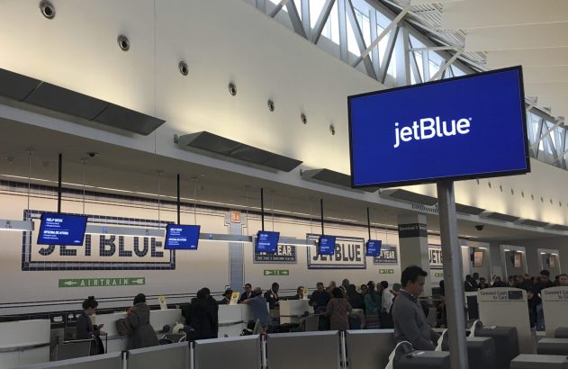 Interior de terminal de aeroporto JetBlue com pessoas em balcões de atendimento e telões exibindo a marca.