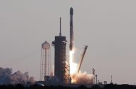 Foguete SpaceX decolando da plataforma, emitindo fortes chamas e fumaça, com uma torre d'água 'SPACE' à esquerda.
