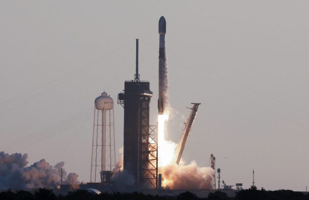 Foguete SpaceX decolando da plataforma, emitindo fortes chamas e fumaça, com uma torre d'água 'SPACE' à esquerda.