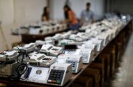 Urnas eletrônicas da Justiça Eleitoral em teste, dispostas em mesas, com técnicos ao fundo.