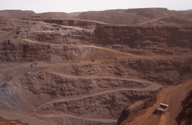 Mina a céu aberto em tons terrosos com caminhões de mineração em seus níveis, um levantando poeira.