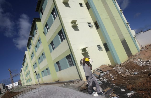 Homem com capacete amarelo carrega saco em obra de prédios residenciais.