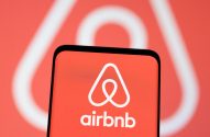 Tela de smartphone com logo e nome Airbnb em fundo vermelho. Ao fundo, logo Airbnb maior e desfocado.