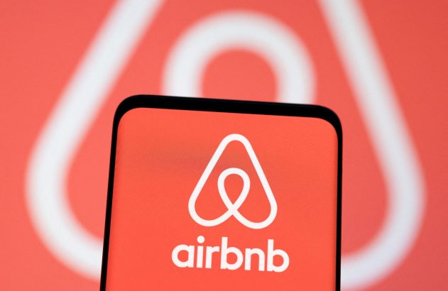 Tela de smartphone com logo e nome Airbnb em fundo vermelho. Ao fundo, logo Airbnb maior e desfocado.