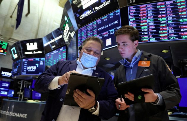 Dois operadores da bolsa, um usando máscara, analisam dados em tablets. Monitores com cotações de ações da NYSE ao fundo.