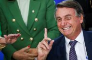 Jair Bolsonaro sorridente, gesticulando com o dedo indicador. Ao fundo, pessoa de blazer verde.