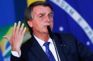 Jair Bolsonaro discursa com a mão levantada e microfone, tendo a bandeira do Brasil como fundo.