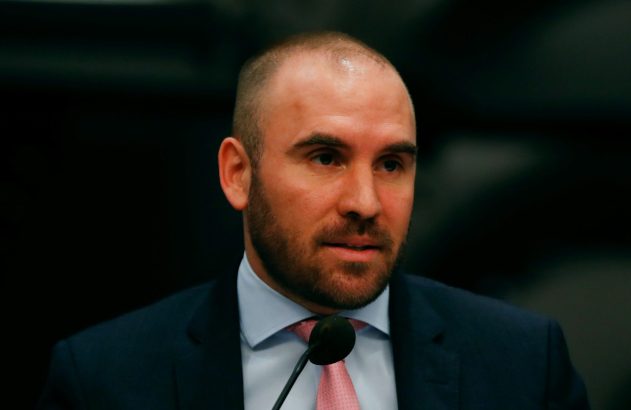 Homem com barba e cabelo curto, de terno escuro e gravata rosa, discursando em um microfone. Fundo desfocado.