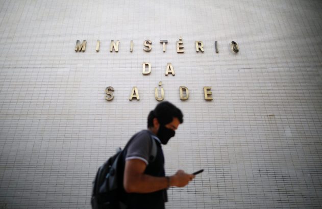 Homem de máscara caminha e olha celular, letreiro "Ministério da Saúde" em parede clara ao fundo.