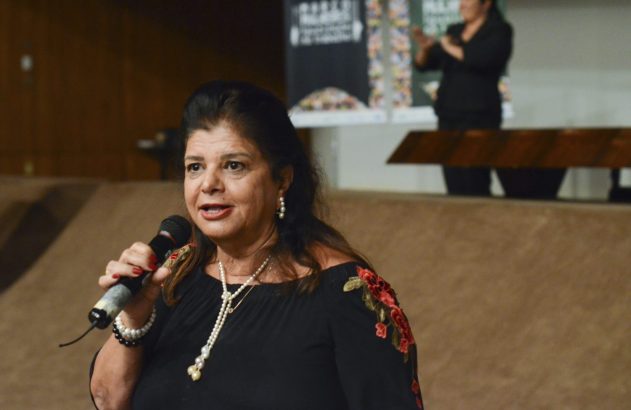 Mulher com blusa preta bordada de flores e colar de pérolas fala em microfone durante um evento.