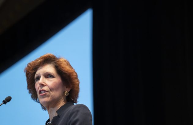 Mulher de cabelos ruivos fala ao microfone contra fundo azul e escuro.