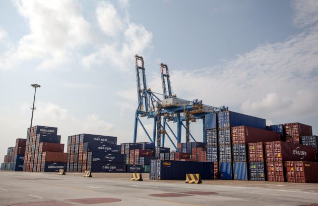 Guindastes e pilhas de contêineres CMA CGM e Seaco em um pátio de terminal portuário.
