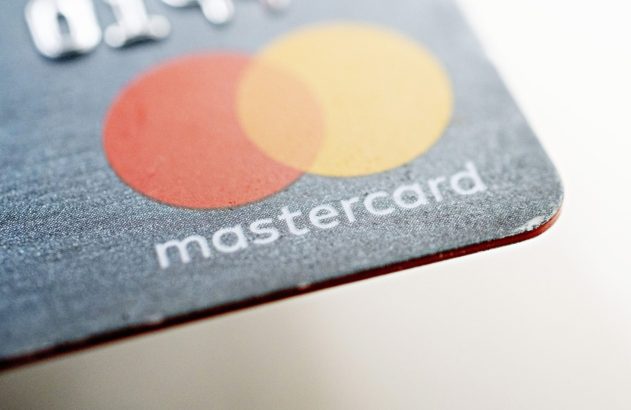 Close-up de um cartão Mastercard cinza com textura. Detalhe do logo vermelho/amarelo e nome 'mastercard', borda vermelha.