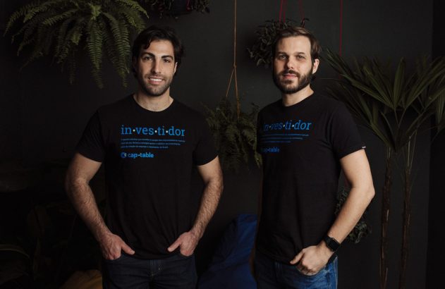 Dois homens sorrindo, vestindo camisetas pretas com "in-ves-ti-dor" e "cap-table". Posam em ambiente interno com plantas.