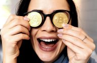 Mulher sorrindo, com moedas douradas de Bitcoin no lugar das lentes dos óculos, boca aberta.