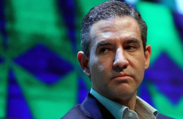 Retrato de um homem em terno azul e camisa listrada, com expressão séria, olhando para a direita. Fundo abstrato verde e azul.