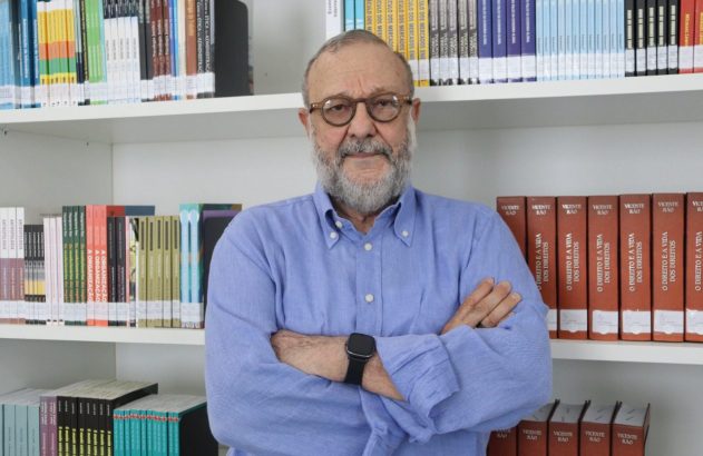 Homem com barba e óculos, camisa azul, braços cruzados, em frente a uma estante de livros.